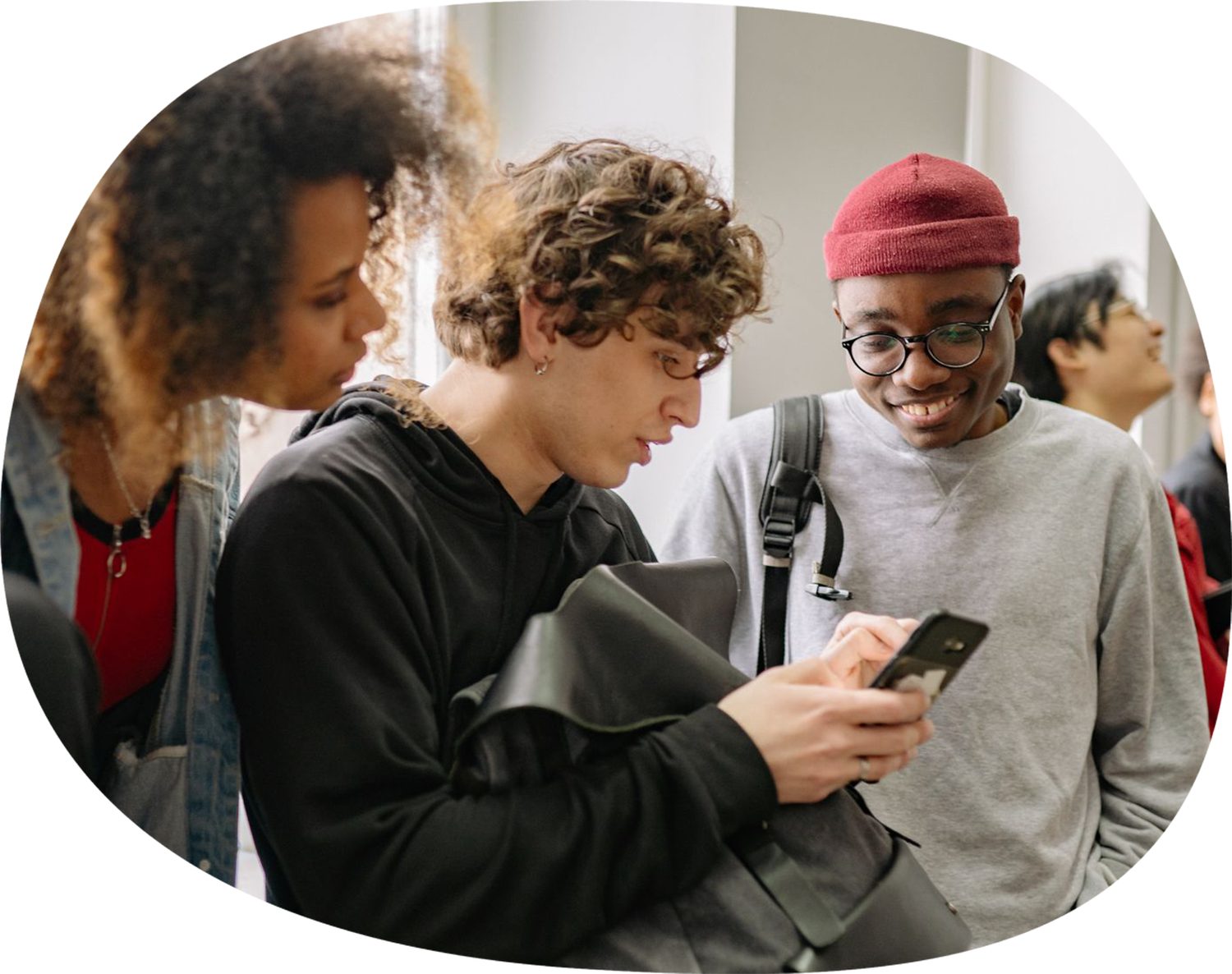 Cinq étudiants réunis dans un couloir vitré ; l'un d'eux montre l'écran de son téléphone à deux camarades penchés vers lui, deux autres discutent en arrière-plan.